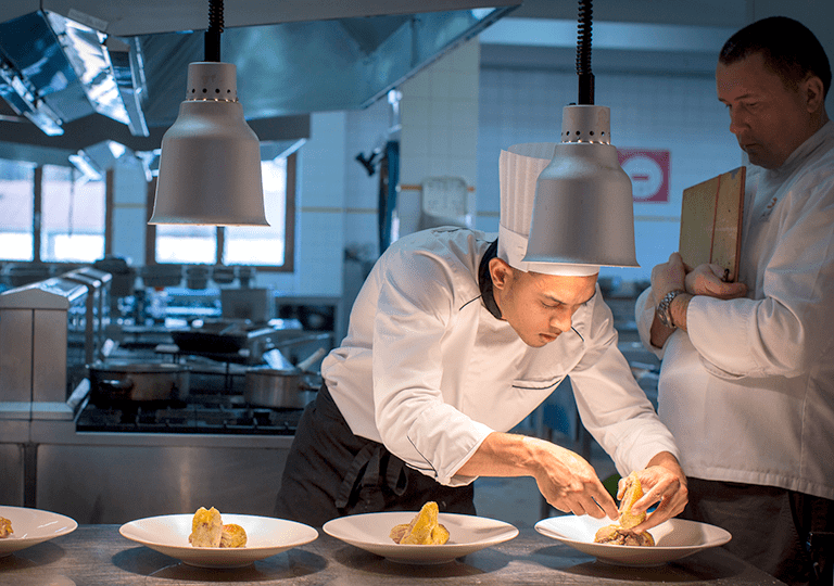 apprenti cuisinier en train de dresser des plats sous les lampes chauffantes devant son professeur à l'école des métiers